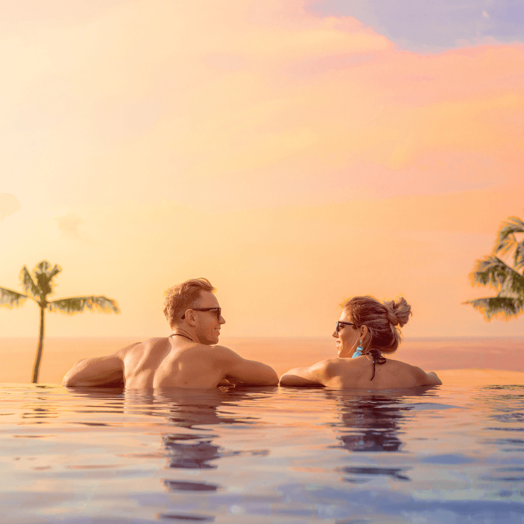 Couple relaxing in an infinity pool at sunset with palm trees in the background.