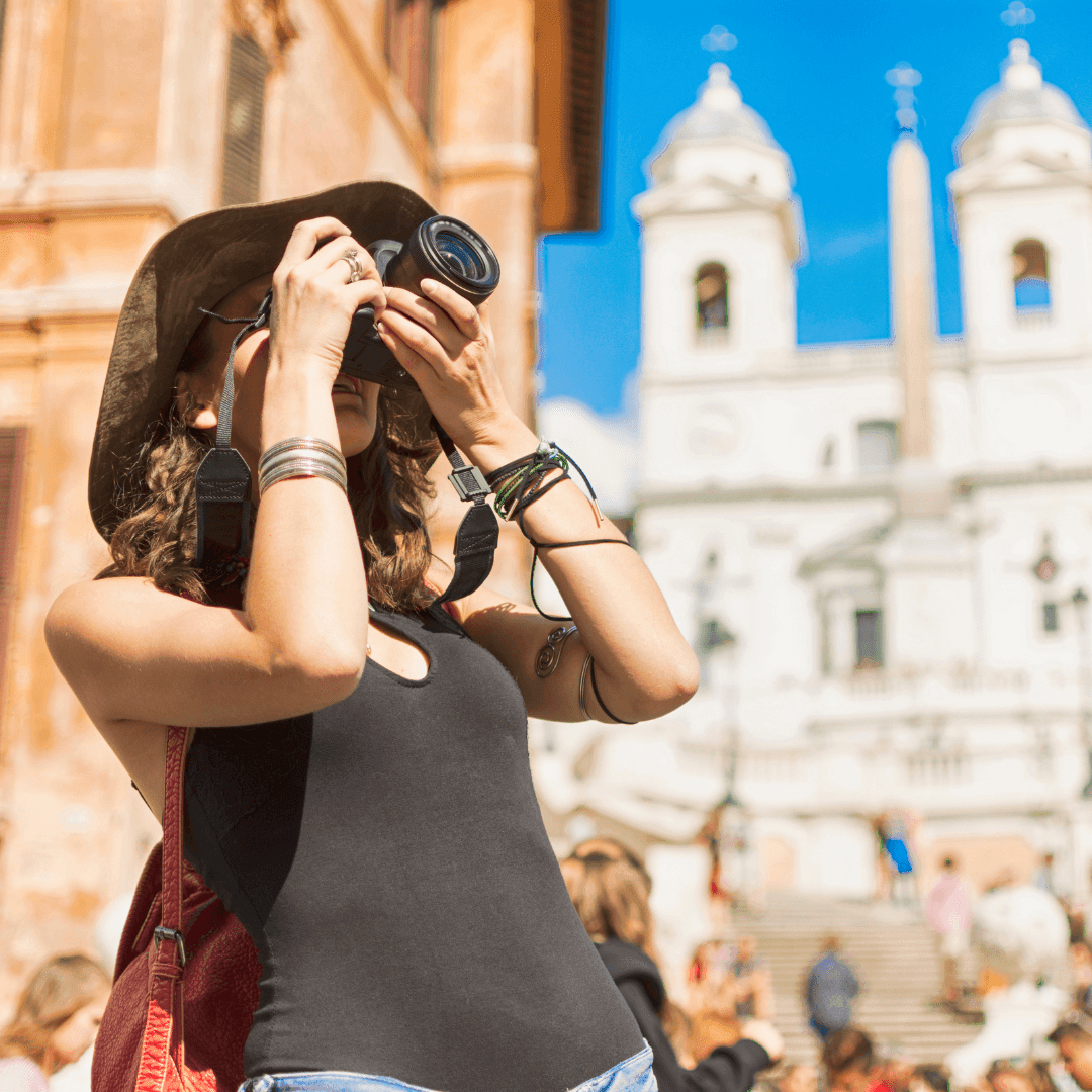 Woman in a hat taking photos in a sunny plaza with historic architecture in the background.