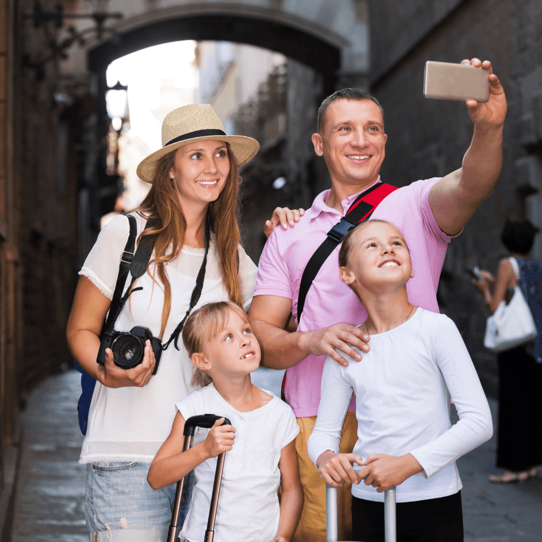 Family taking a selfie in a narrow historic street, holding luggage and camera.